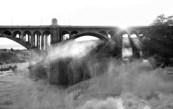 A black and white image of a bridge over a river with water splashing against it.