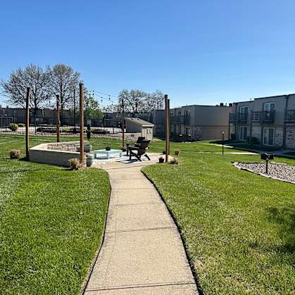 A walkway through a park with benches and buildings in the background, offering a peaceful picnic area at Park 25 Apartments.