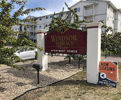 The exterior sign with trees and surrounded by greenery with the buildings in the background of Windsor Terrace Apartment Homes. 