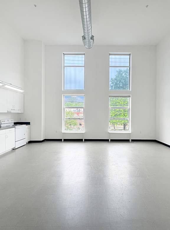 A large empty room with a kitchen area to the left and a window with a view of the outside.