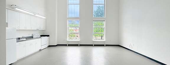A large empty room with a kitchen area to the left and a window with a view of the outside.
