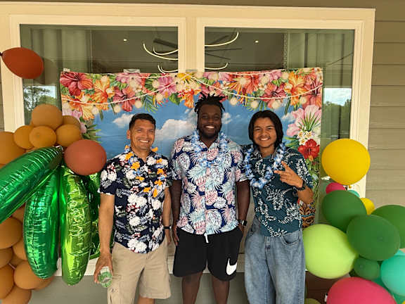 Three people wearing floral shirts stand in front of a balloon arch.