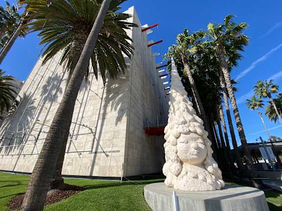 A sculpture of a face is in front of a building with palm trees.