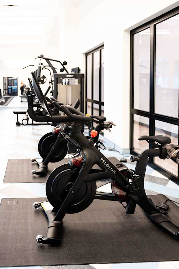 A Peloton bike is on a mat in a brightly lit gym.