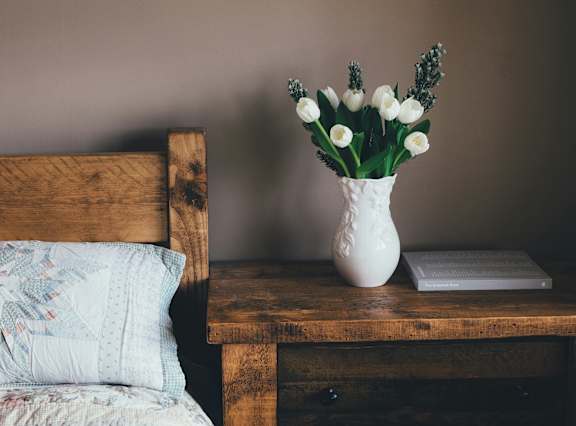 a white vase with flowers on a table next to a bed