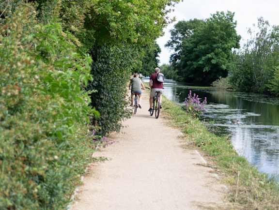 Two people riding bikes on a path next to a river. at The Woodlands Apartments, Milton Keynes, MK5 6GS