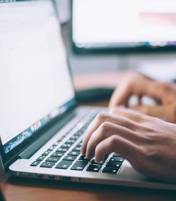 A person is typing on a laptop with a monitor in the background.