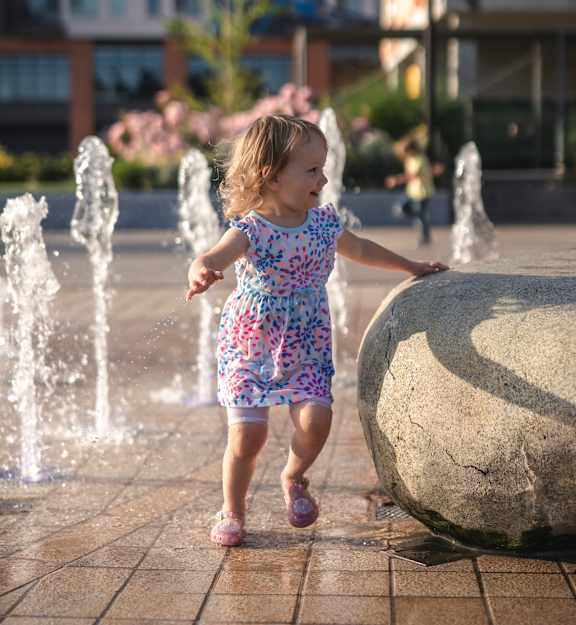 A little girl in a floral dress is playing with a large rock in front of a fountain.