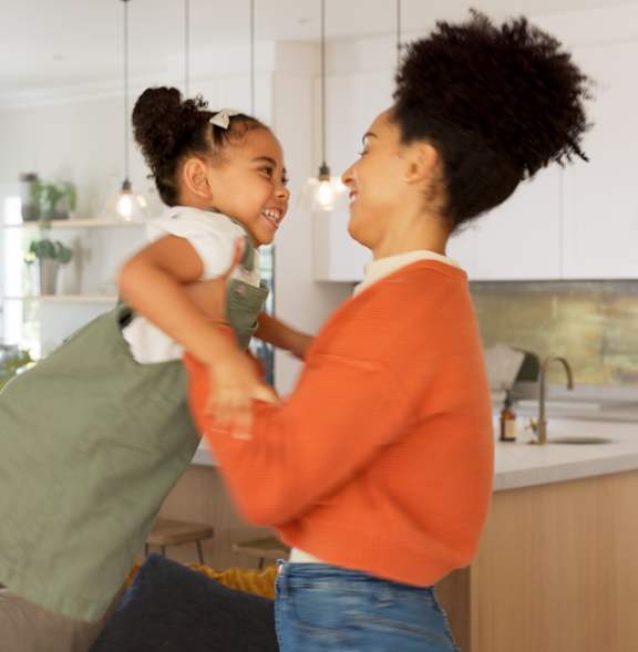 A woman in an orange top is holding a child in a green dress in a kitchen.