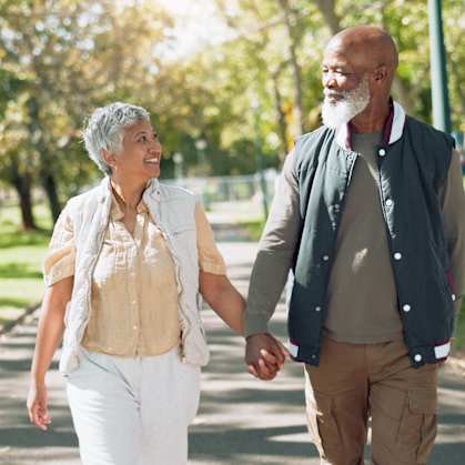 An elderly couple is walking hand in hand in a park.