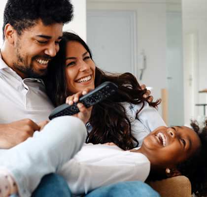 A family of three is sitting on a couch, with the woman holding a remote control.