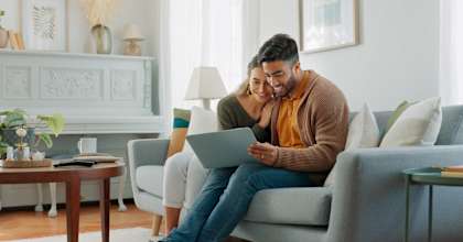 A man and woman are sitting on a couch, with the man holding a tablet and the woman looking at it lovingly.