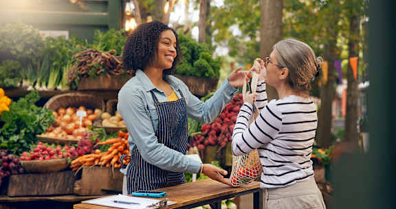 A woman in a striped shirt is buying vegetables from a woman in an apron at a market.