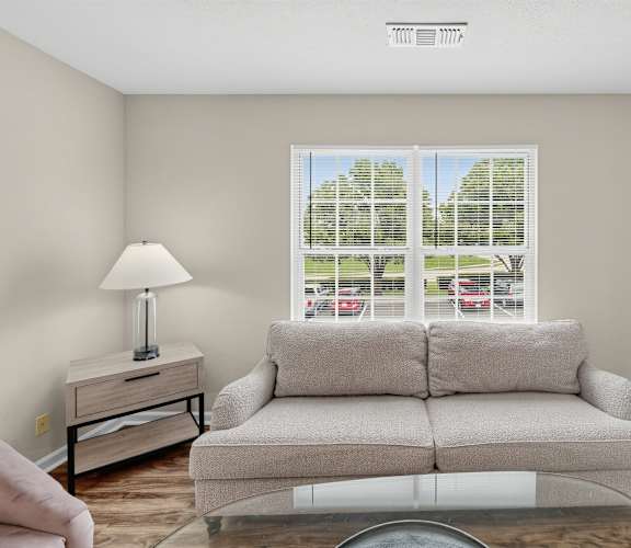 A living room with a grey couch, a lamp, and a window with a view of a parking lot.