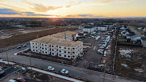 A large building under construction is surrounded by a parking lot and other buildings.