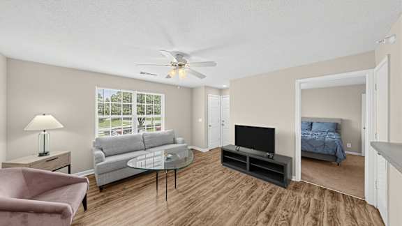 A living room with a pink chair, a grey couch, a glass coffee table, a flat screen TV, and a bed with blue bedding.
