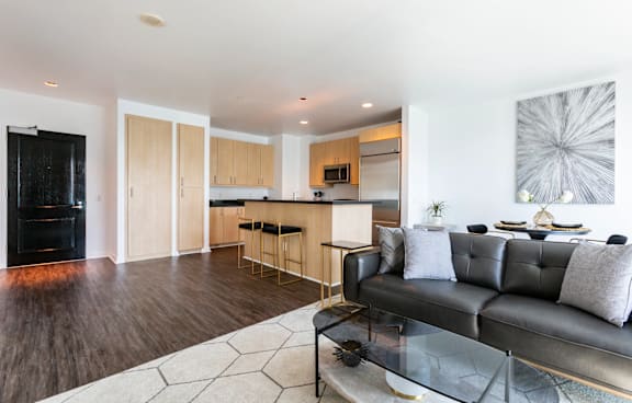 A black couch sits in a living room with a wooden floor and a bar area in the background.
