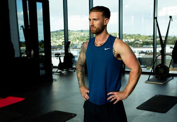 A man with tattoos stands in a gym wearing a blue Nike tank top.