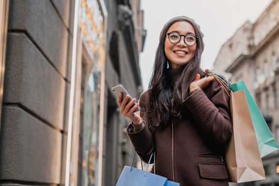 A woman with glasses and a brown coat is holding shopping bags and looking at her phone. at Trio Newton Apartments, Newton, 02460