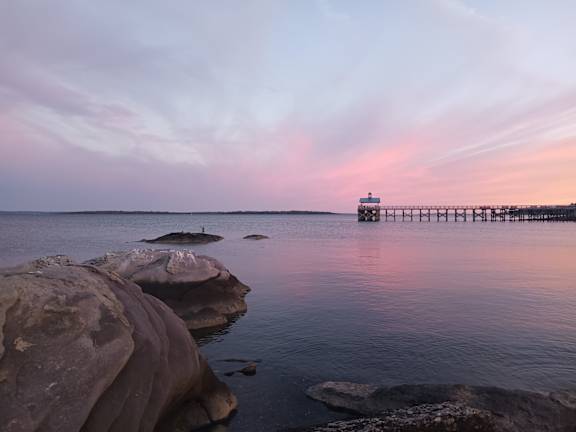 A long pier extends into a calm body of water with rocks in the foreground.