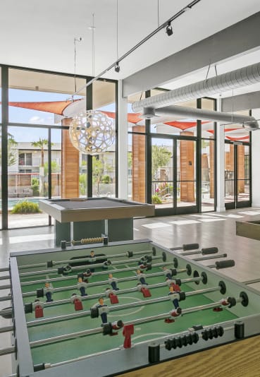 A modern game room at Sentry Apartments offers foosball, pool, and shuffleboard tables, with large windows overlooking the pool.