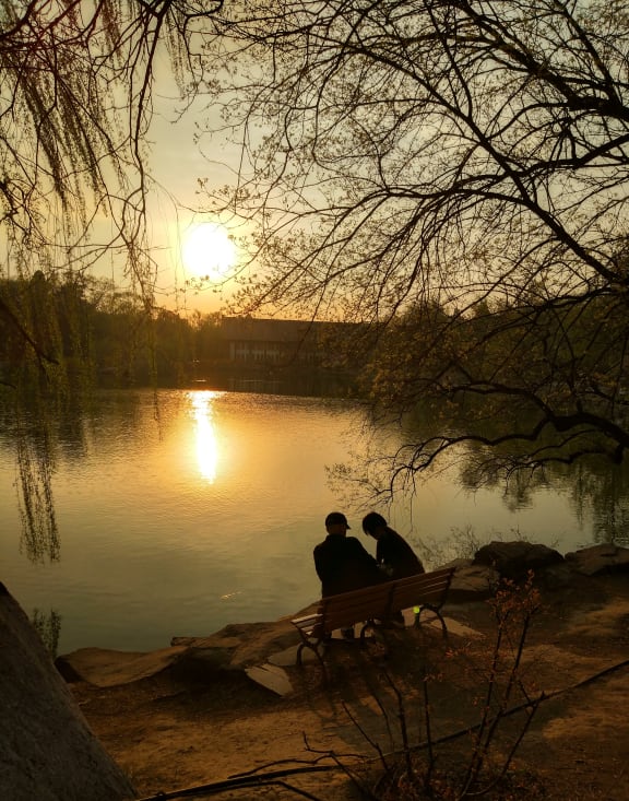 two people sitting on a bench overlooking a lake at sunset