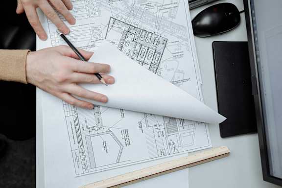 A person is working on architectural drawings with a computer monitor, keyboard, and mouse in the background.