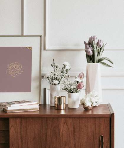 A framed picture with a gold design is on a table with flowers and a book.