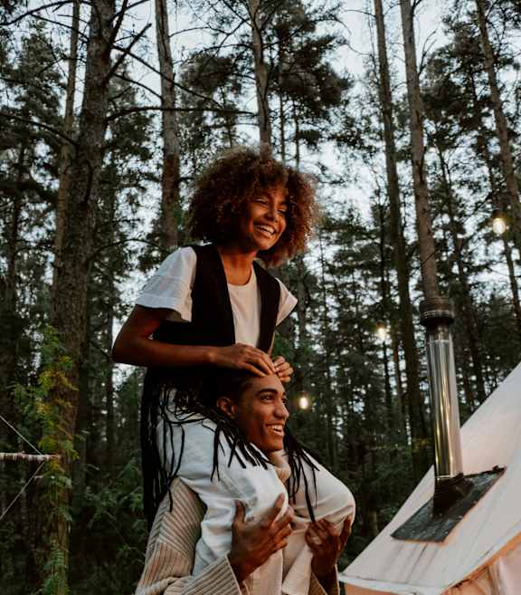 A person with dreadlocks is standing on a wooden platform in a forest, holding a book and wearing a sweater and jeans.