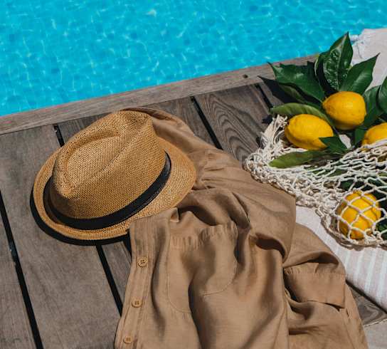 A hat, a jacket, and a bag of lemons are on a wooden table by a pool.
