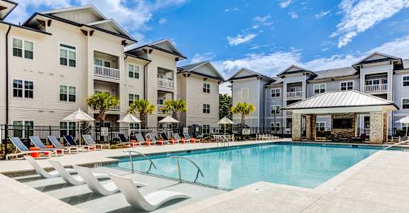 A swimming pool surrounded by lounge chairs and umbrellas in front of apartment buildings.