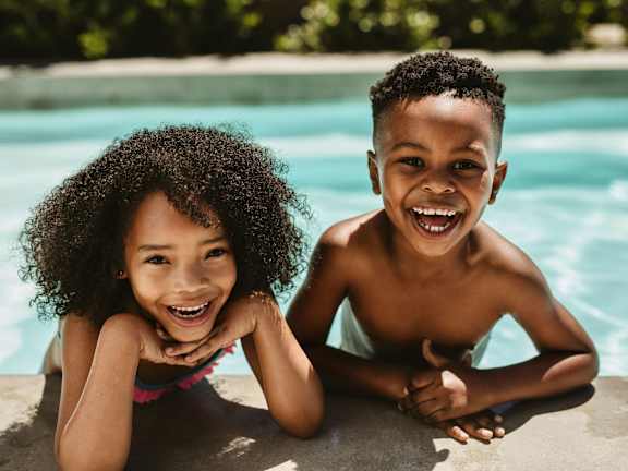 Two children are smiling and sitting by a pool.