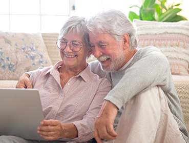 An older couple is sitting on a couch and looking at a laptop together.