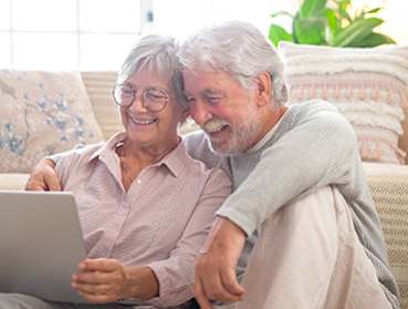 An older couple is sitting on a couch and looking at a laptop together.