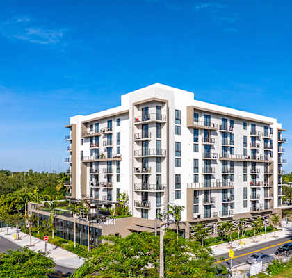A large white apartment building with balconies is surrounded by green trees and a clear blue sky.