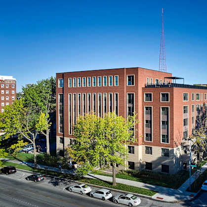 A large red building with a tall antenna on the roof is surrounded by trees and cars.
