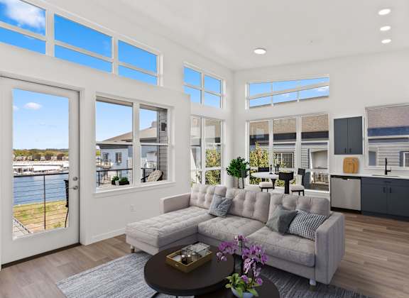 A modern living room with a large grey sofa and a coffee table in front of a glass wall overlooking a waterfront.