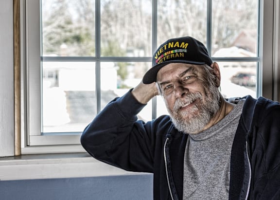 A man wearing a Vietnam veteran hat sits by a window.