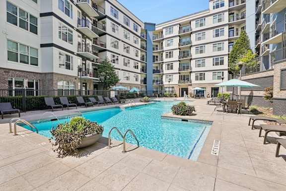 A pool surrounded by chairs and umbrellas in front of apartment buildings.