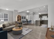 Thumbnail 3 of 4 - A modern living room with a grey sofa, a wooden coffee table, and a dining area in the background.