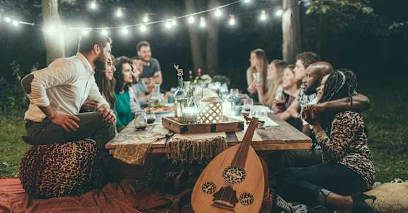 A group of people are sitting around a picnic table in a forest at night, enjoying a meal and each other's company.