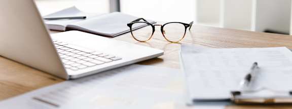 A desk with a laptop, eye glasses and a notebook