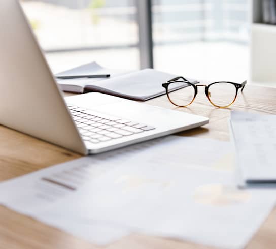 a desk with a laptop, eye glasses and a notebook