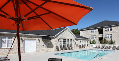 A large orange umbrella is shading a table and chairs by a pool.