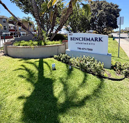 A sign for Benchmark Apartments stands in a grassy area with palm trees.