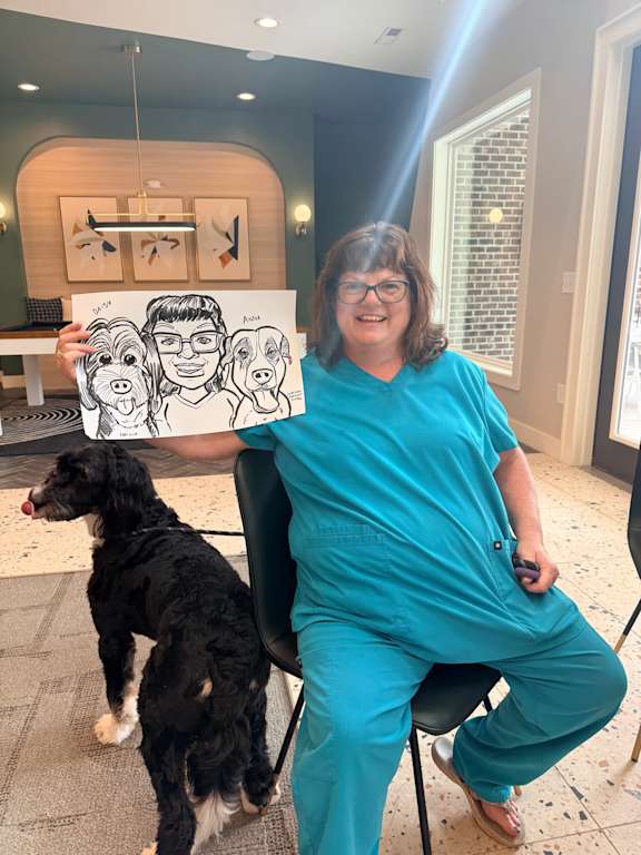 A woman in blue scrubs holds a drawing of a dog while sitting on the floor.