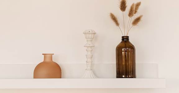 A white shelf with a brown vase, a white candle holder, and a brown bottle with dried plants in it.