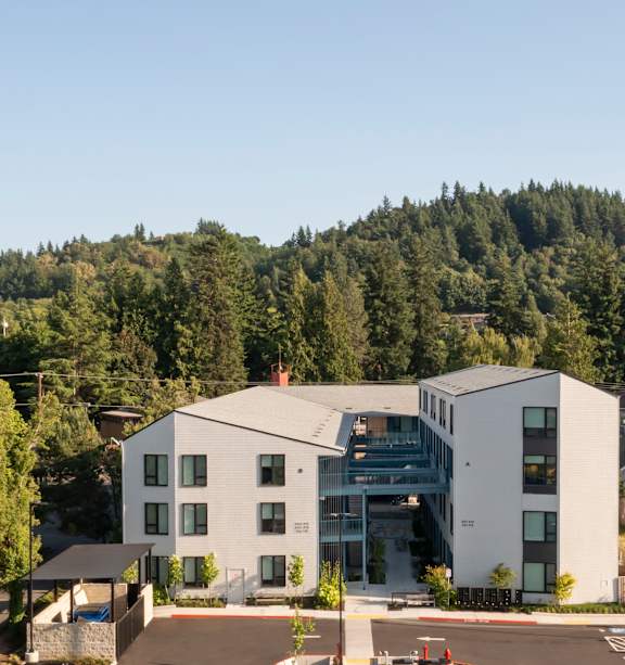A parking lot in front of a large white building with a forest in the background.