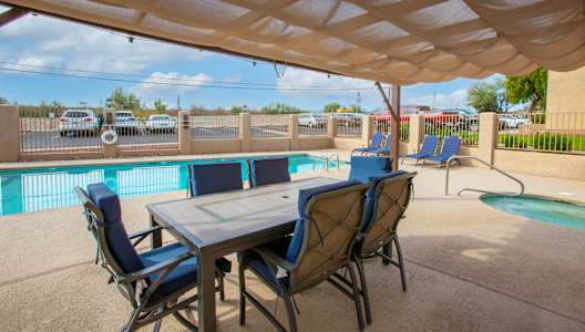 A table with chairs is set up under a canopy by a pool.