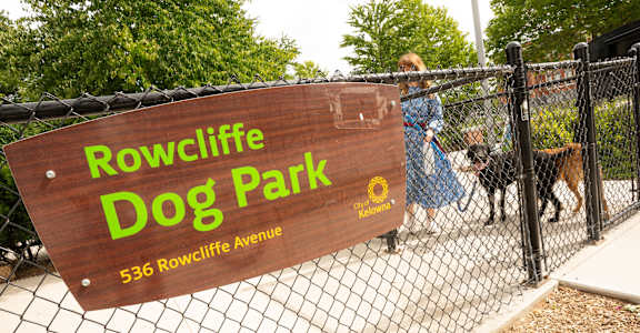 A woman walks her dog in front of a sign that says Rowcliffe Dog Park.